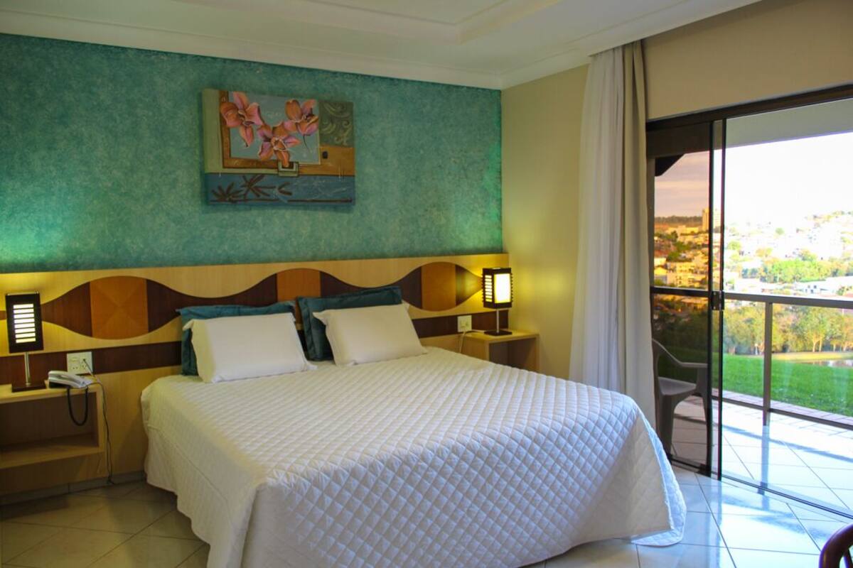 Habitación de hotel con una cama grande, dos mesitas de noche con lámparas, un cuadro en la pared y vistas a un paisaje urbano a través de un ventanal amplio.