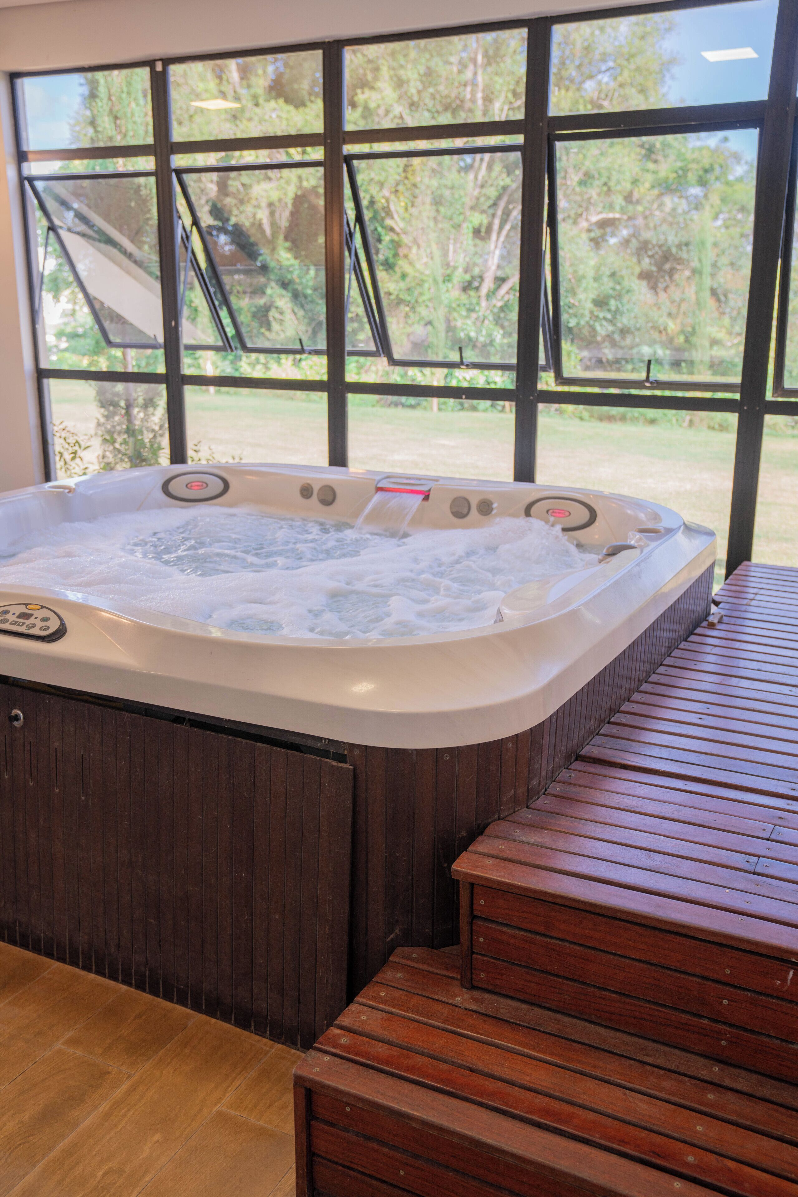 A hot tub with a wooden bench in front, situated indoors with a view of a garden through large windows.