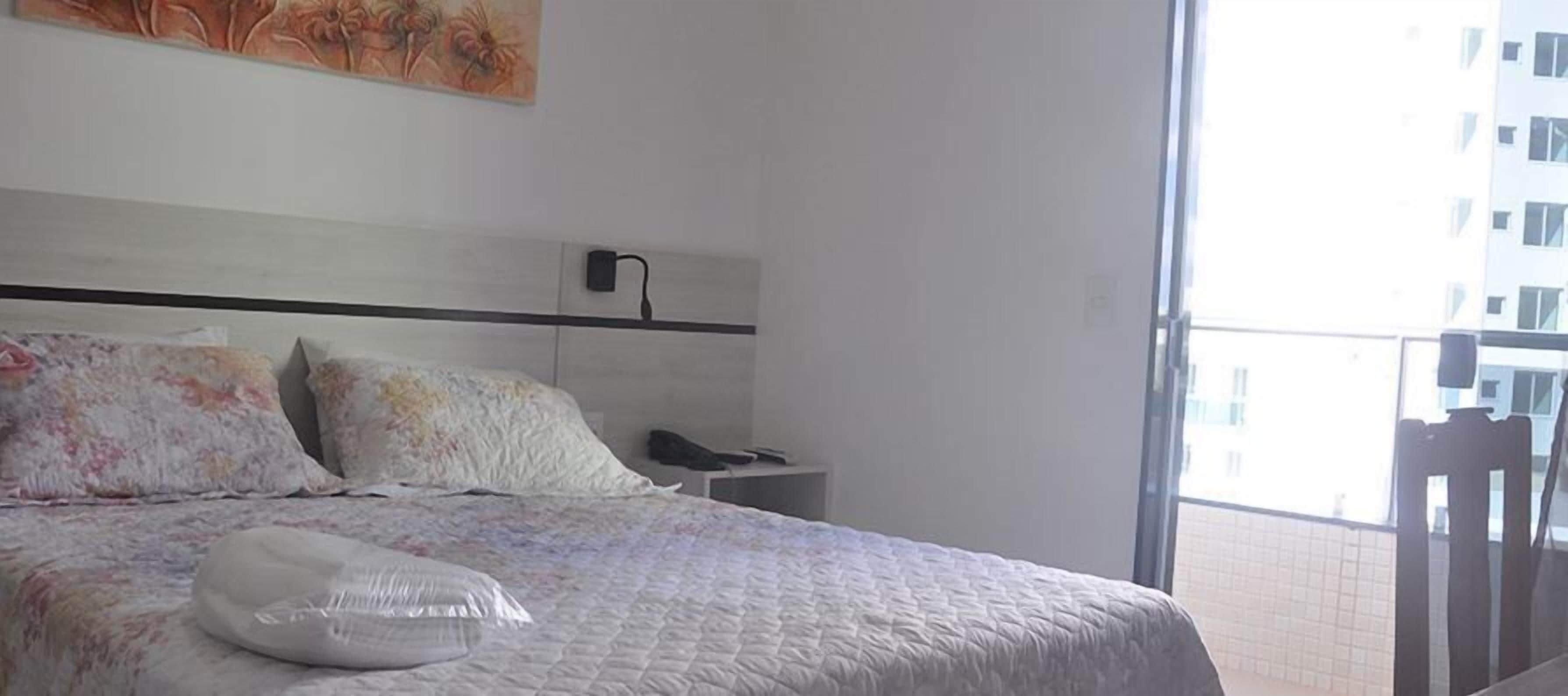 A hotel room with a bed, a nightstand, a wall-mounted lamp, and a window with a view of a building.