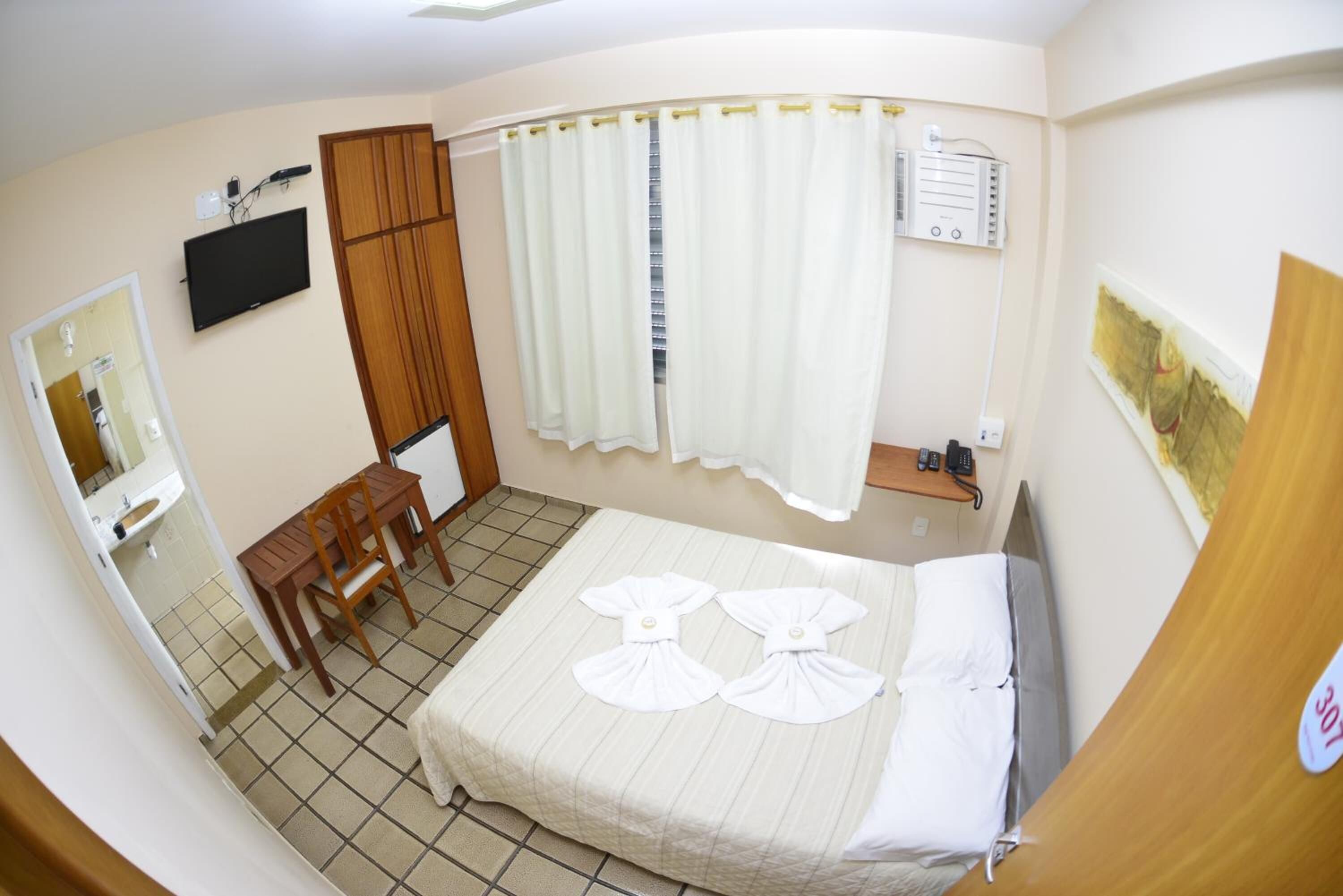 Habitación de hotel con cama, escritorio, silla, televisor, ventana con cortinas y un calentador fijado a la pared.