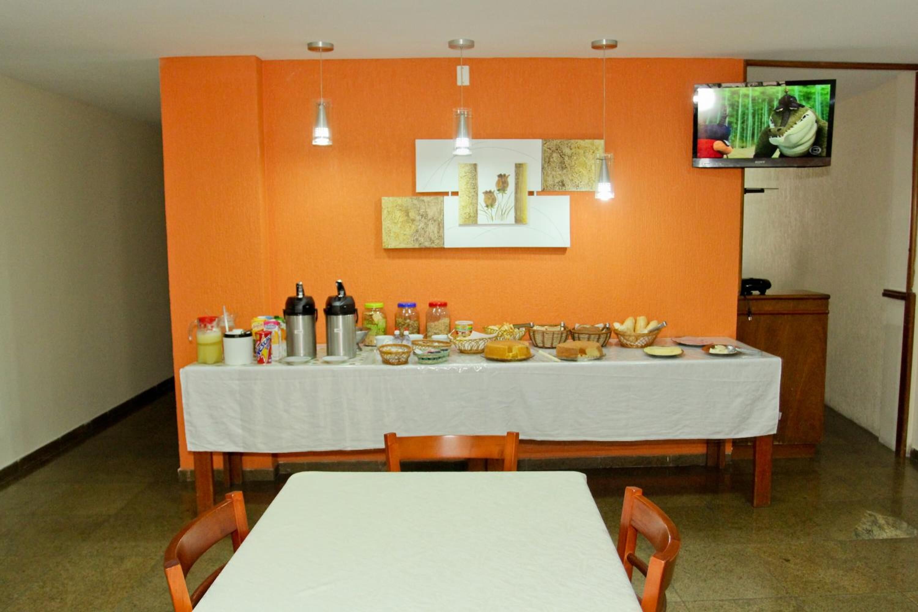 Un comedor con una mesa dispuesta para cuatro, un televisor montado en la pared y diversos alimentos y condimentos sobre la mesa.