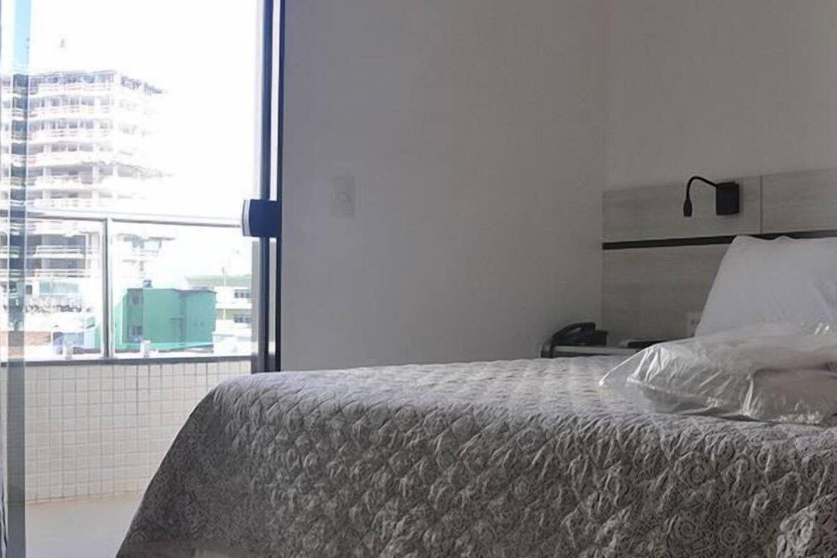 Una habitación de hotel con cama, mesita de noche, teléfono y ventana con vistas a edificios.