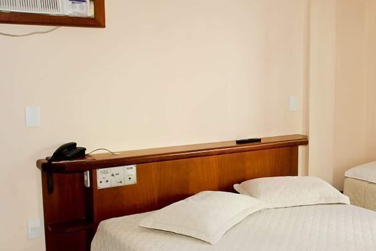Habitación de hotel con dos camas individuales, cabecera de madera, teléfono en la mesita de noche y una ventana con cortinas amarillas.