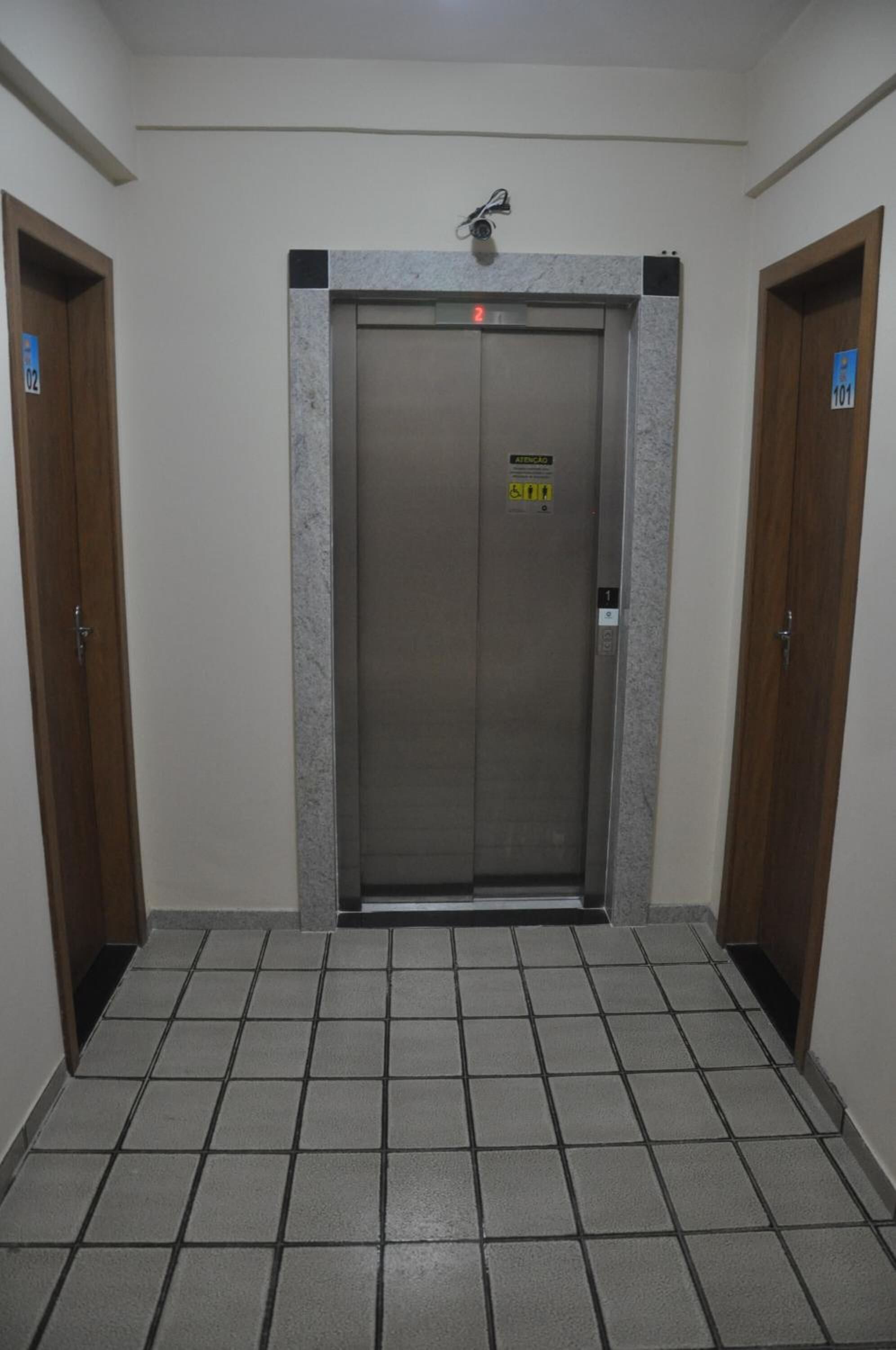 An elevator with a digital display and emergency contact sign, flanked by wooden doors, in a hallway with tiled flooring.