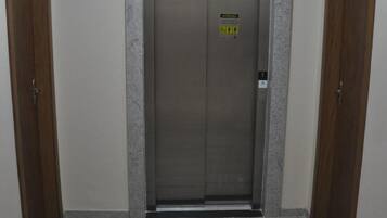 An elevator with a digital display and emergency contact sign, flanked by wooden doors, in a hallway with tiled flooring.