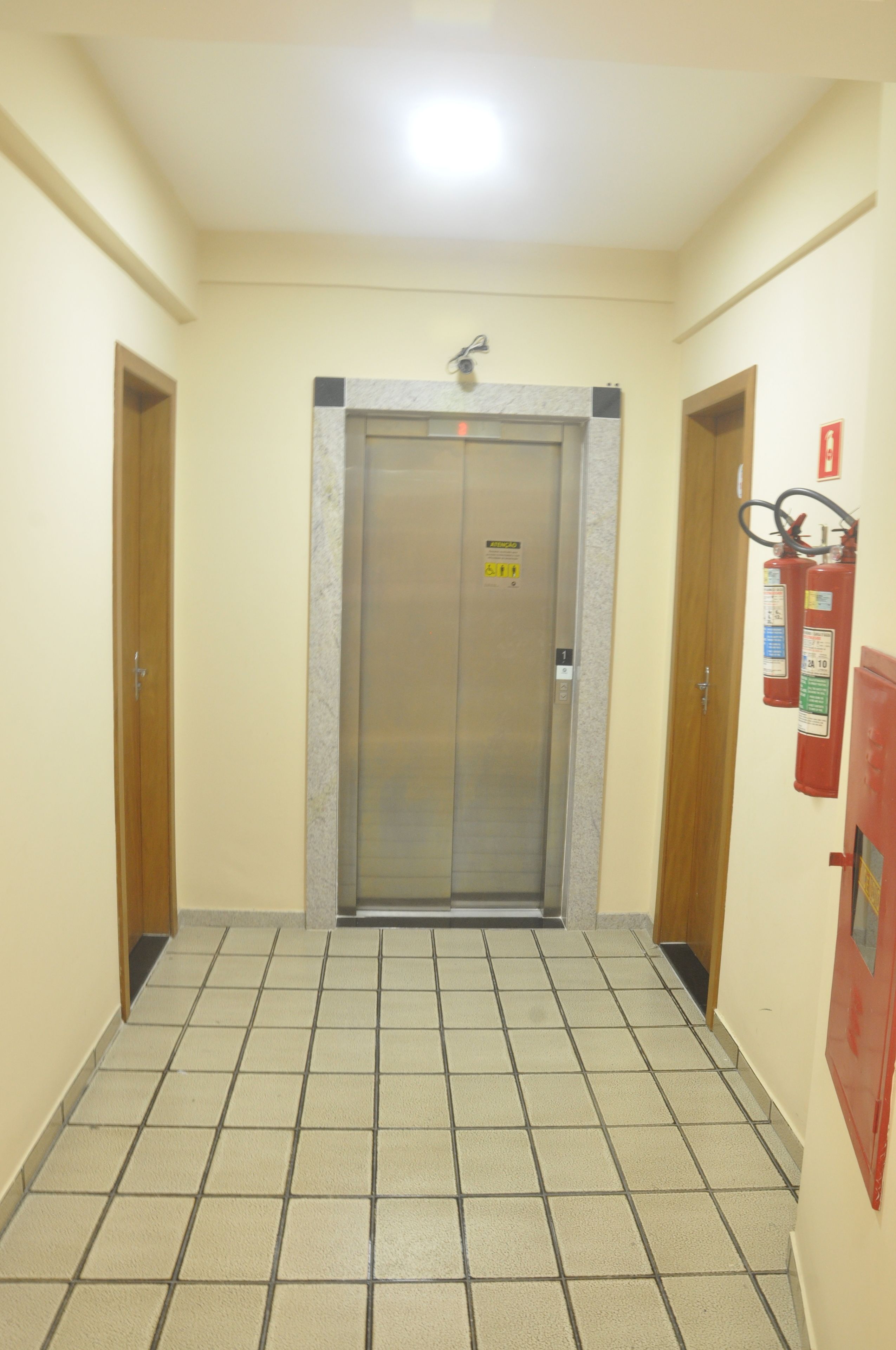 Un couloir avec un sol carrelé, une porte sur laquelle est fixé un extincteur et un luminaire au plafond.