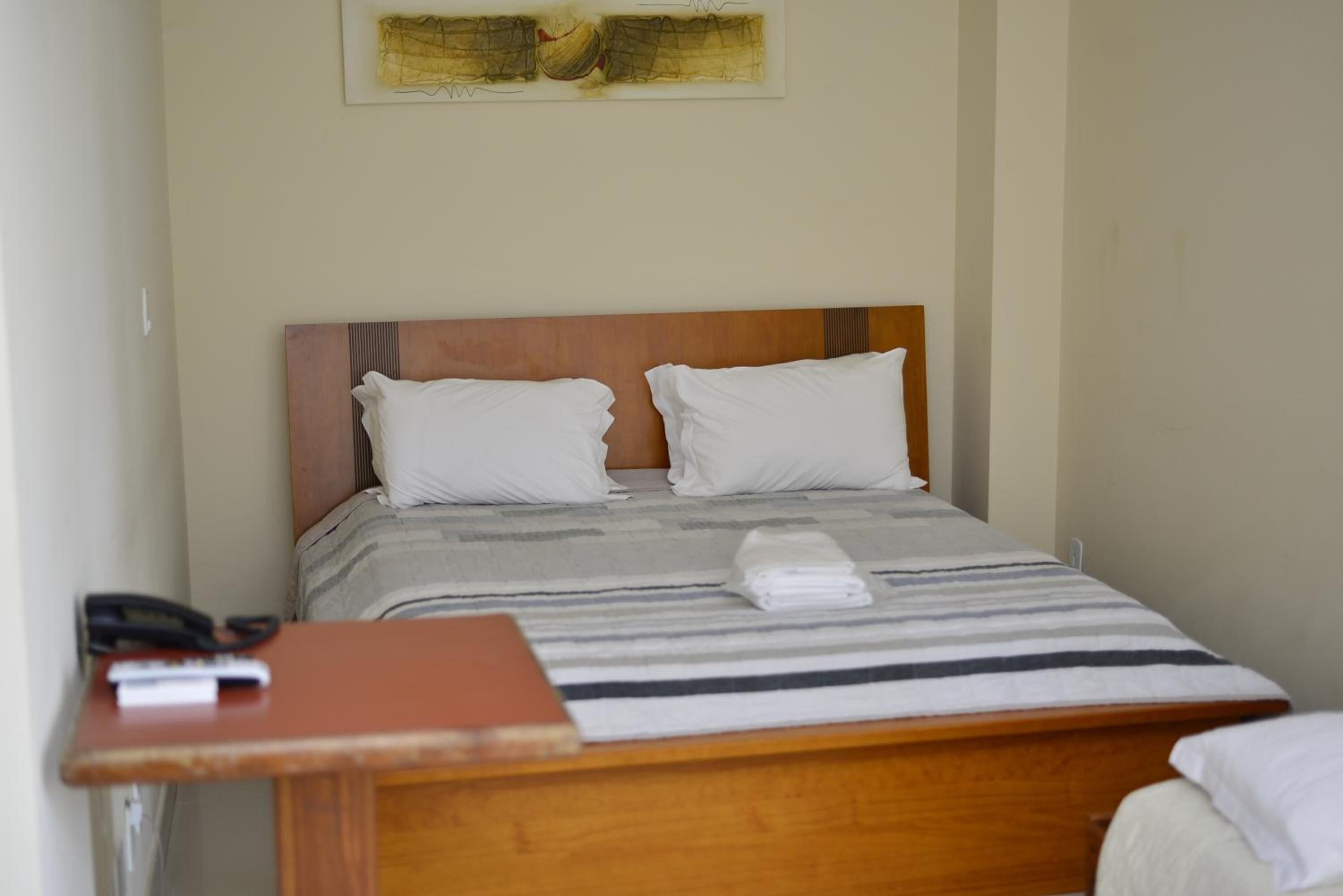 Uma cama de solteiro com cabeceira de madeira, almofadas brancas e um cobertor listrado. Um pequeno criado-mudo com um telefone e um livro está à esquerda.
