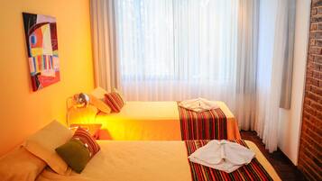 Habitación de hotel con dos camas, un ventanal con cortinas translúcidas, pared de ladrillo y un cuadro abstracto colorido en la pared.