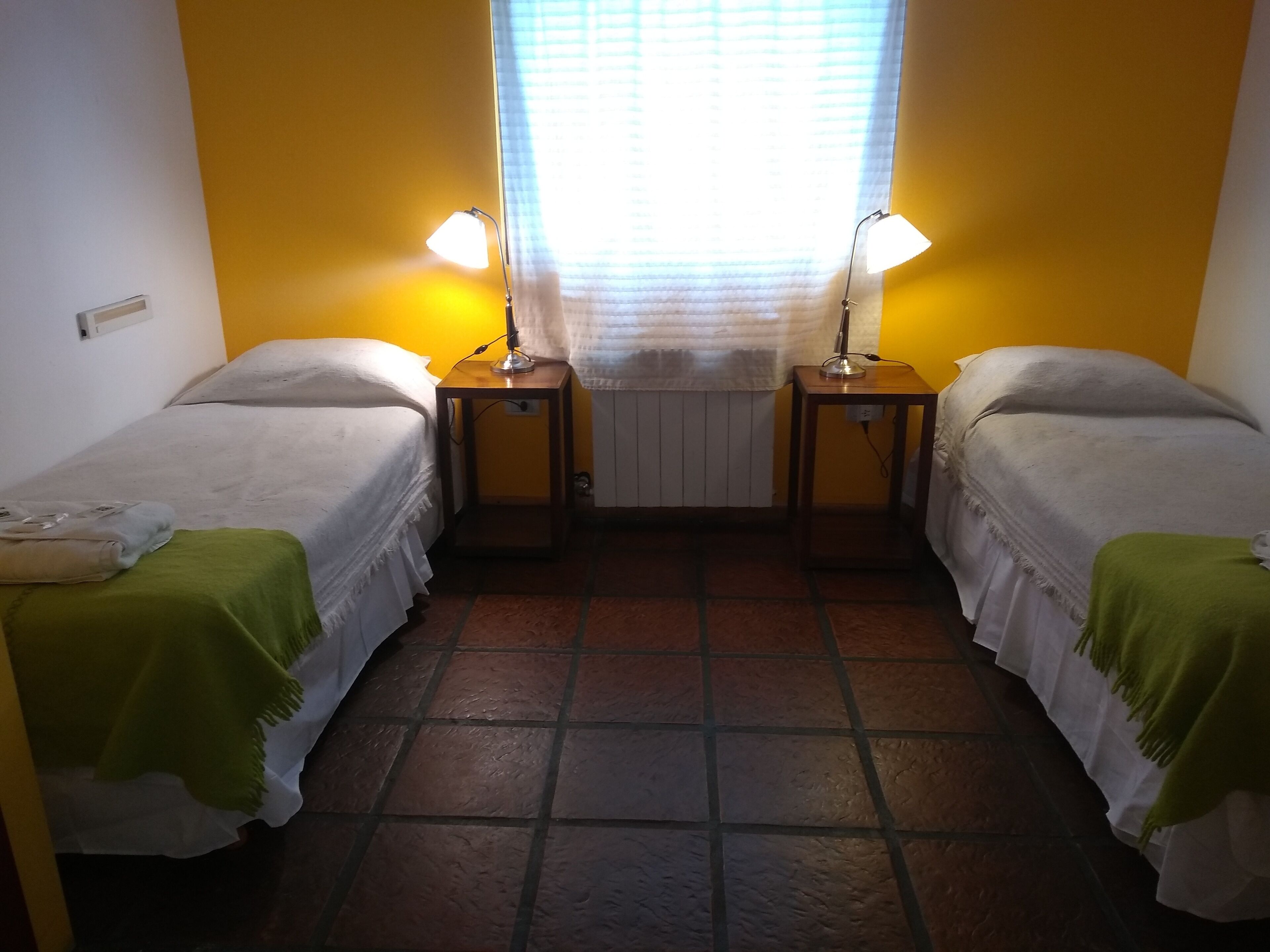 Una habitación pequeña y limpia con dos camas individuales, cada una con una manta verde y una lámpara sobre una mesita de noche de madera.