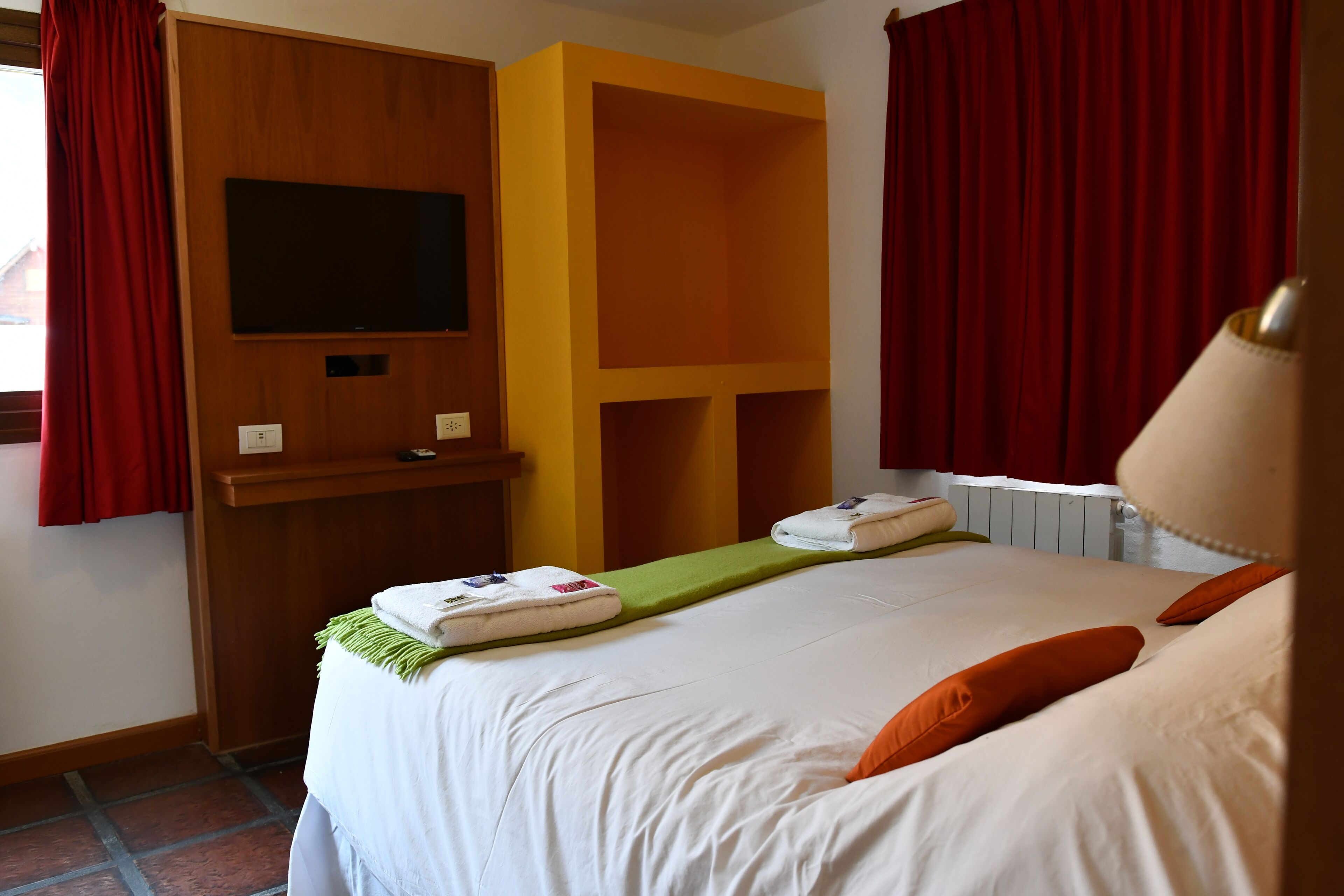 Ein Hotelzimmer mit Bett, einem an einer Holzplatte montierten Fernseher, einer Regaleinheit und roten Vorhängen.