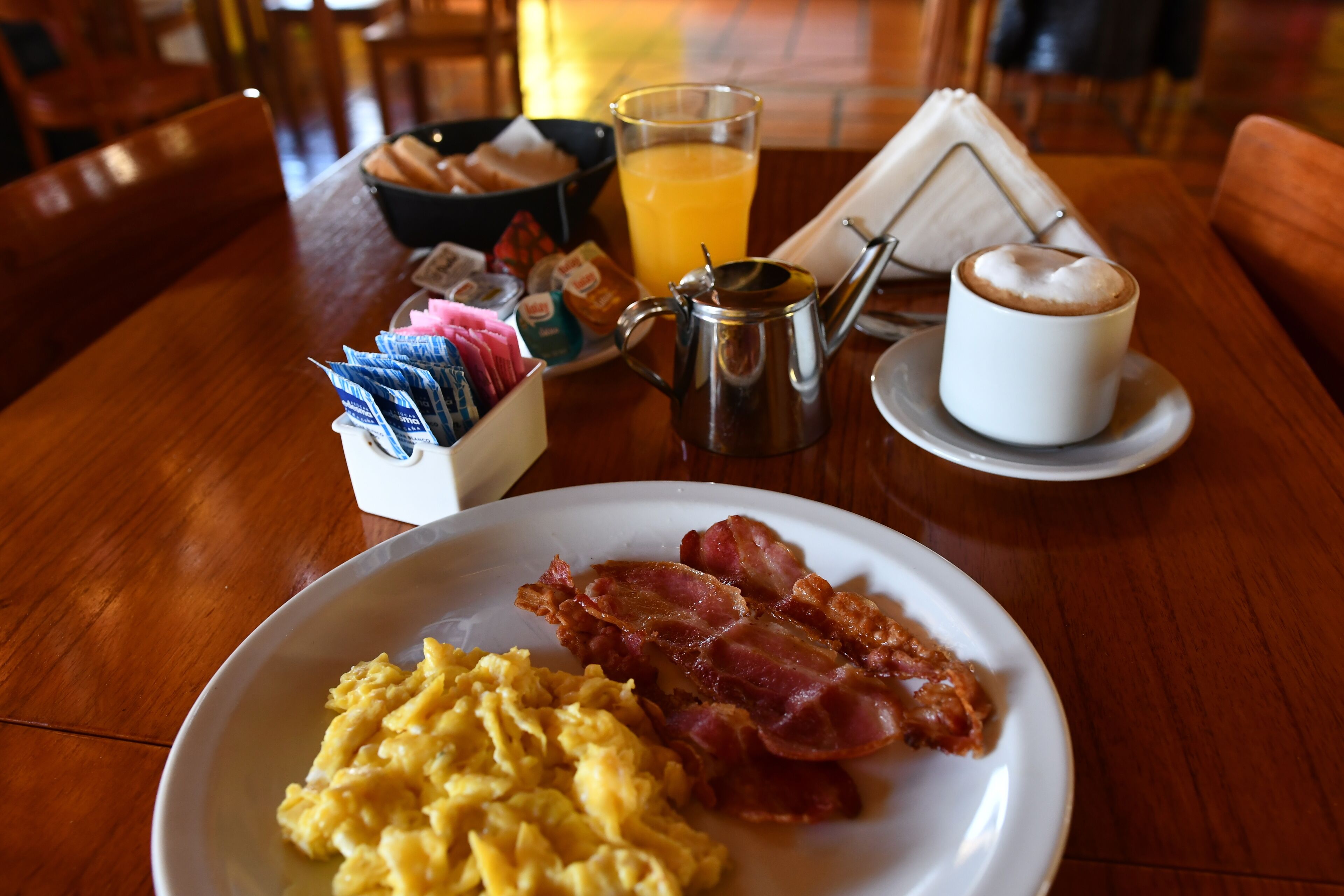 A breakfast table with scrambled eggs, bacon, a coffee cup, a glass of orange juice, and a teapot.