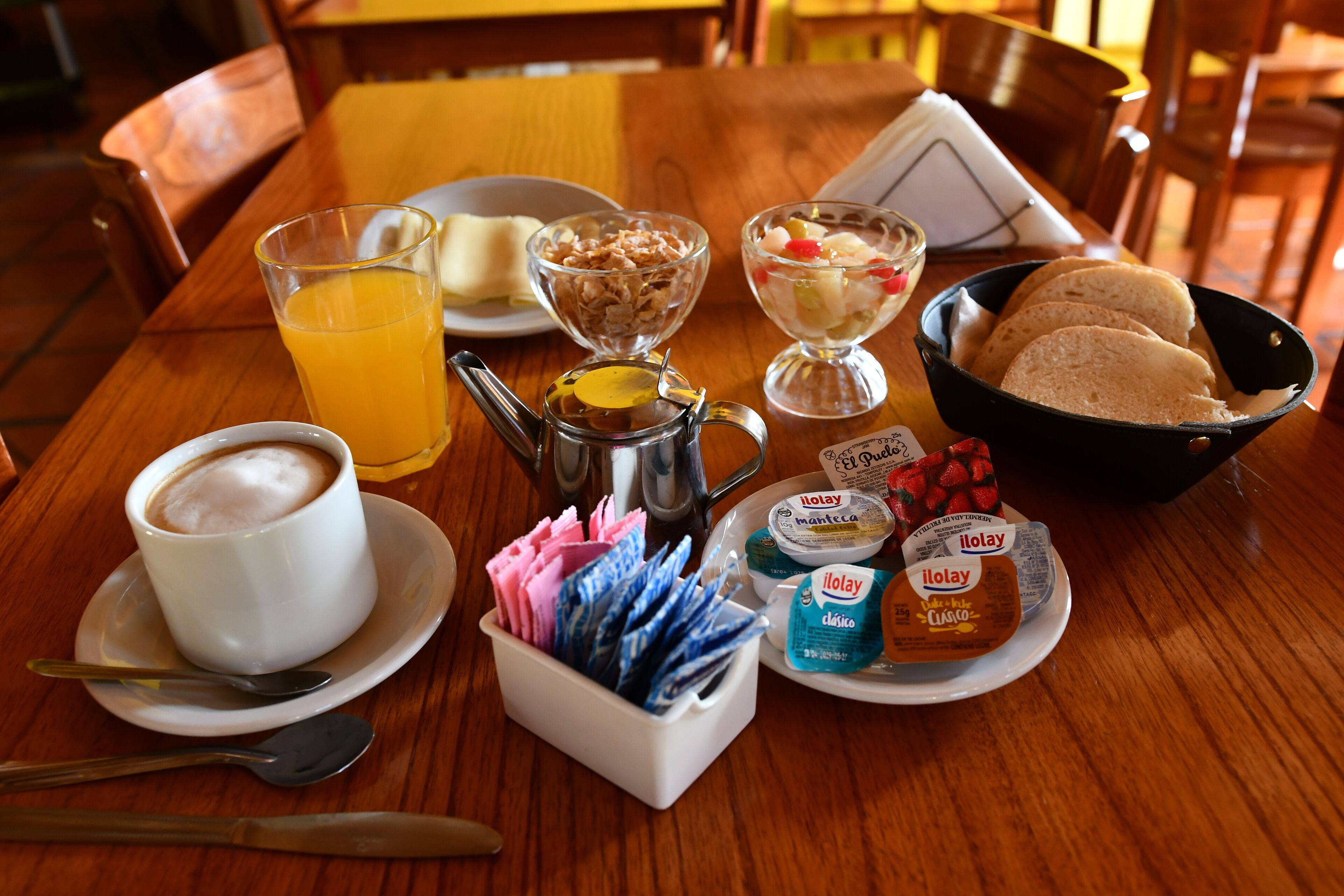 Una mesa de desayuno con una taza de café, un vaso de jugo de naranja, un tazón de cereal, un recipiente pequeño de yogur, un plato de pan y un termo.