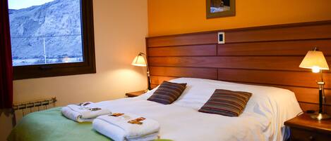 Habitación de hotel con una cama grande, dos almohadas, una manta verde, dos lámparas de noche, una ventana con vistas a la montaña y un cuadro en la pared.
