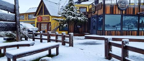 A snow-covered outdoor area with wooden benches and a hostel with a yellow facade.
