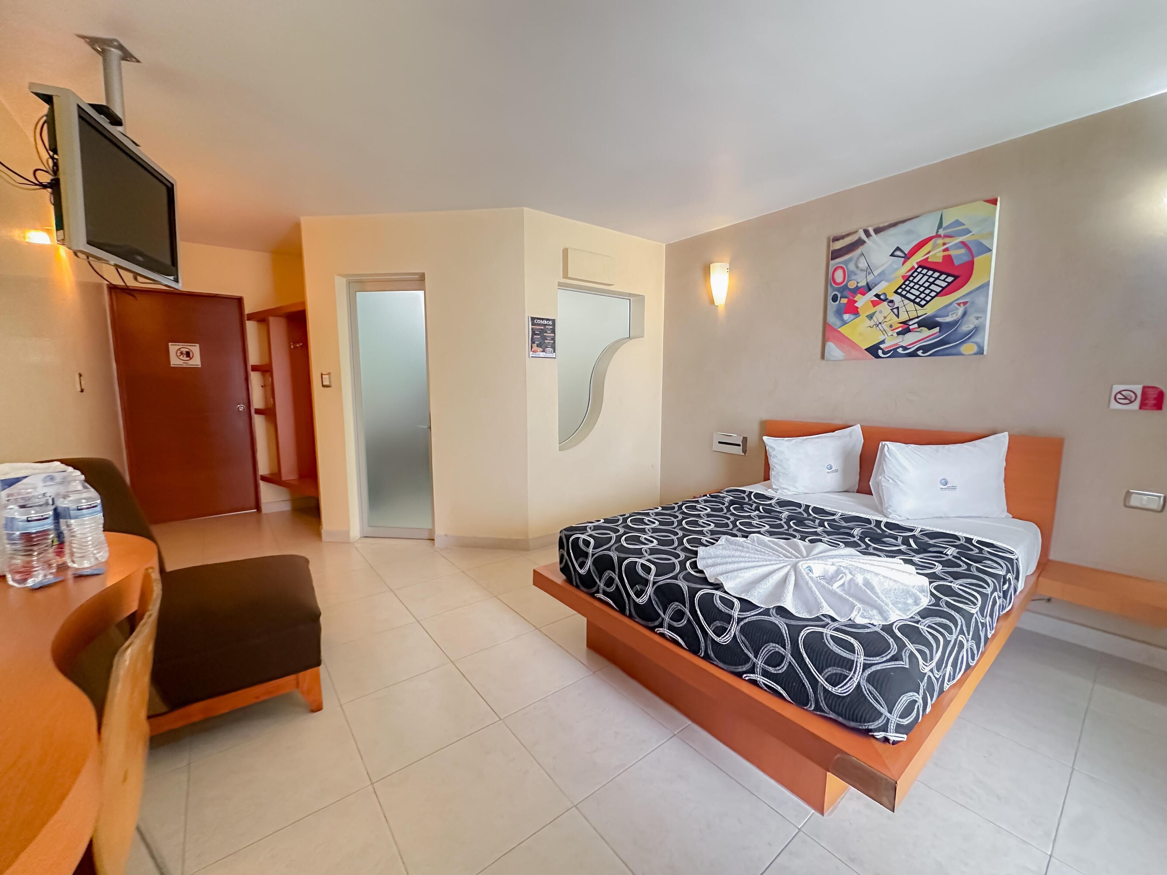 A hotel room with a bed, a desk, a television, and a painting on the wall.