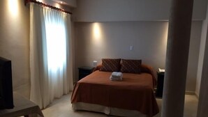 A hotel room with a bed, a nightstand, a table, and a window with curtains. - Hotel San Carlos (Termas de Rio Hondo)