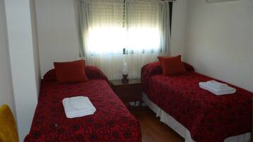 Une chambre avec deux lits, chacun recouvert d’une couvre-lit à motifs rouges et agrémenté d’oreillers blancs. On y trouve une fenêtre avec des rideaux légers, une petite table et un climatiseur fixé au mur.