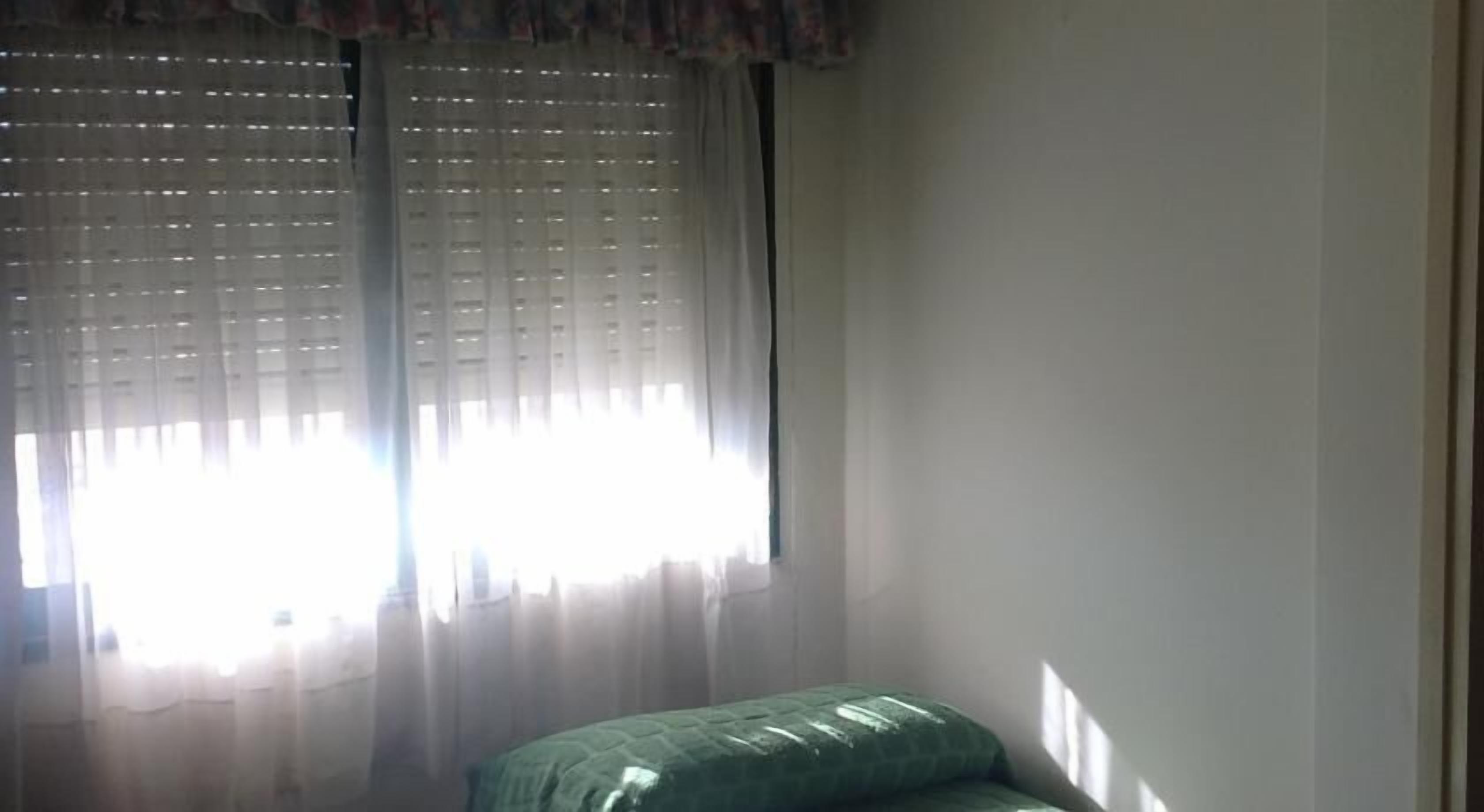 A room with a bed, a window with sheer curtains, and a wall.