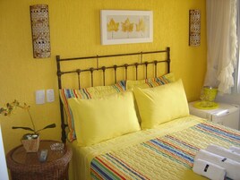 Un dormitorio de color amarillo con cama, almohadas, una mesita de noche, una lámpara y una obra de arte colgada en la pared.