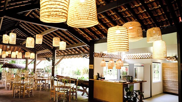 A restaurant with wooden tables and chairs, hanging wicker lamps, and a bar area with a counter and refrigerator.