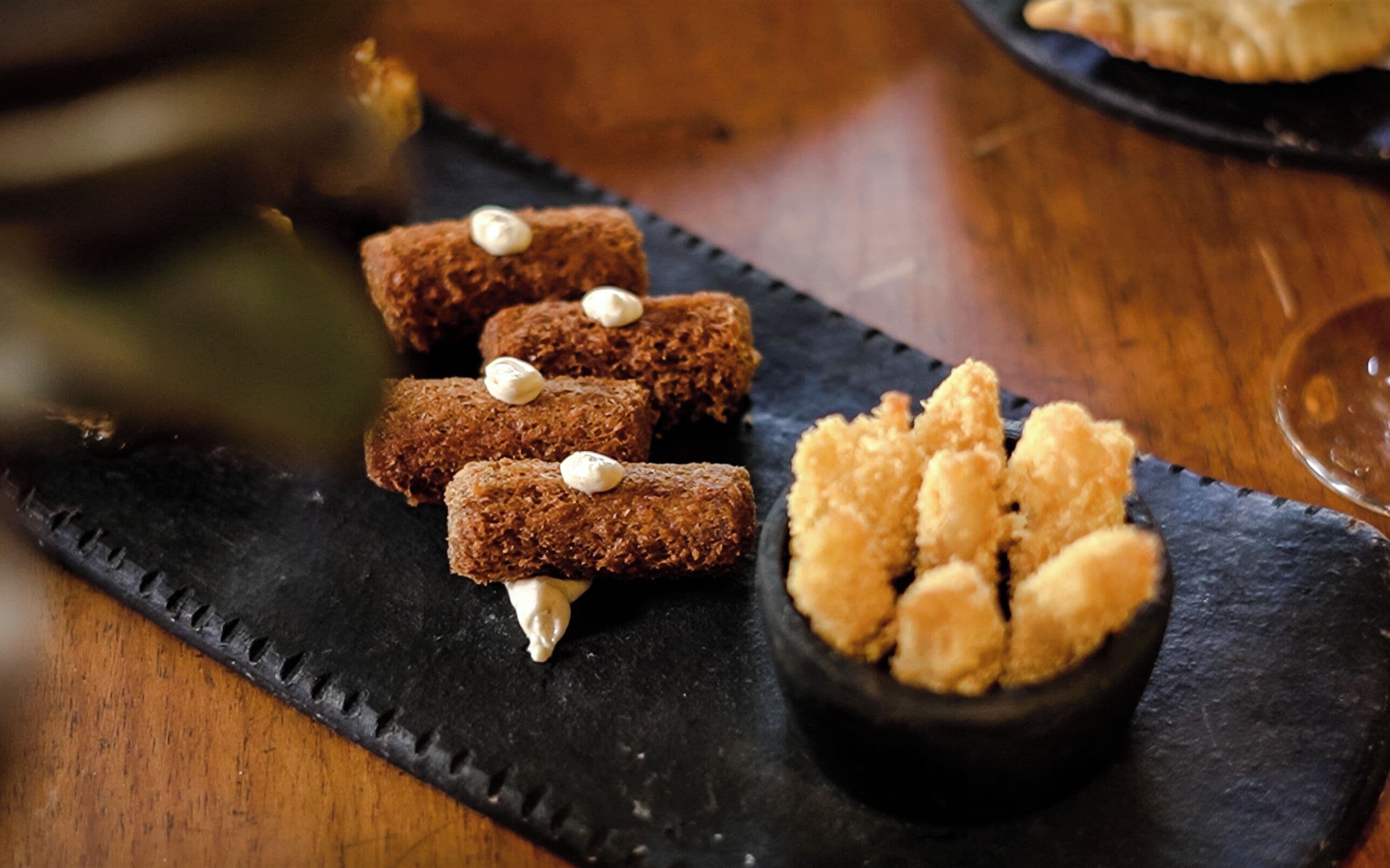 Un plato de comida con tres elementos cilíndricos marrones cubiertos por una sustancia blanca, y un bol pequeño con porciones fritas doradas.