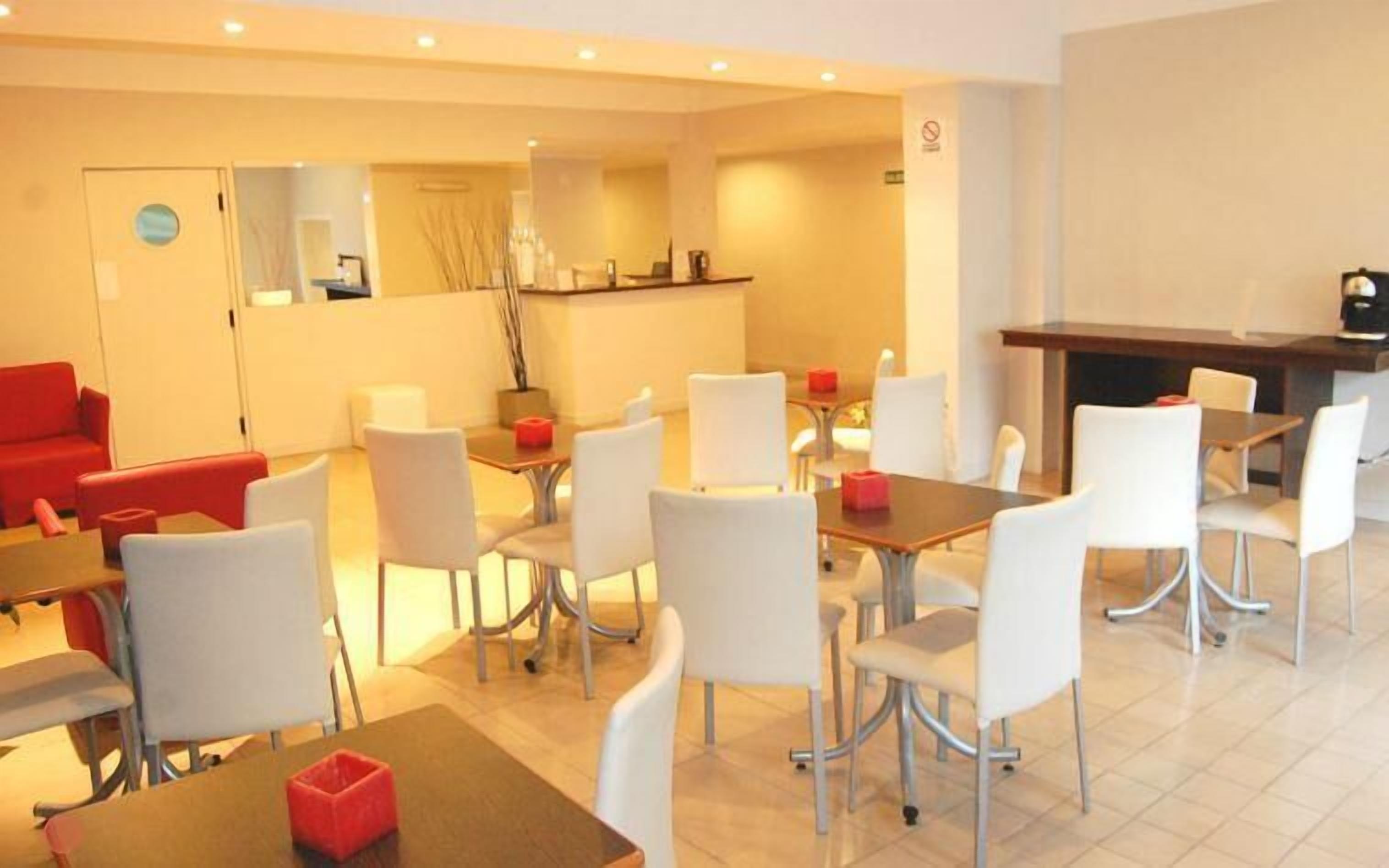 A modern hotel dining area with white chairs, red cushions, and a central bar area.