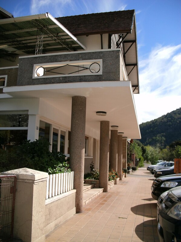 A modern building with a covered walkway, stone columns, and a row of parked cars. - Hotel Crismalu (San Martín de los Andes)