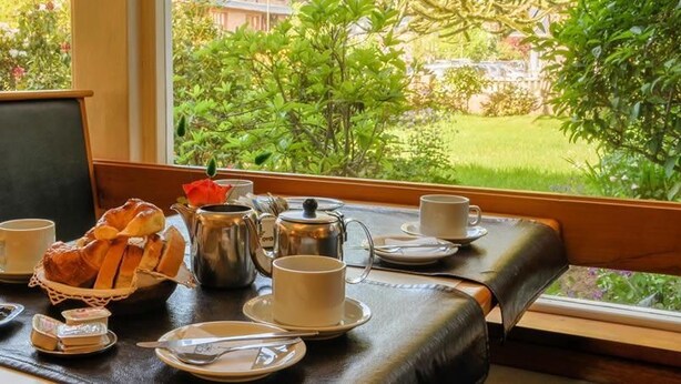A table set with a teapot, cups, plates, and a basket of bread, with a view of greenery outside. - Hotel Crismalu (San Martín de los Andes)