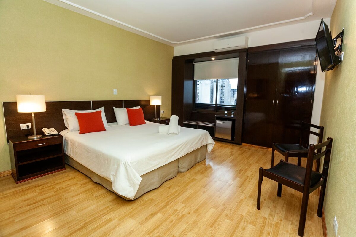 Habitación de hotel con cama, mesitas de noche, escritorio, silla, televisor y armario.