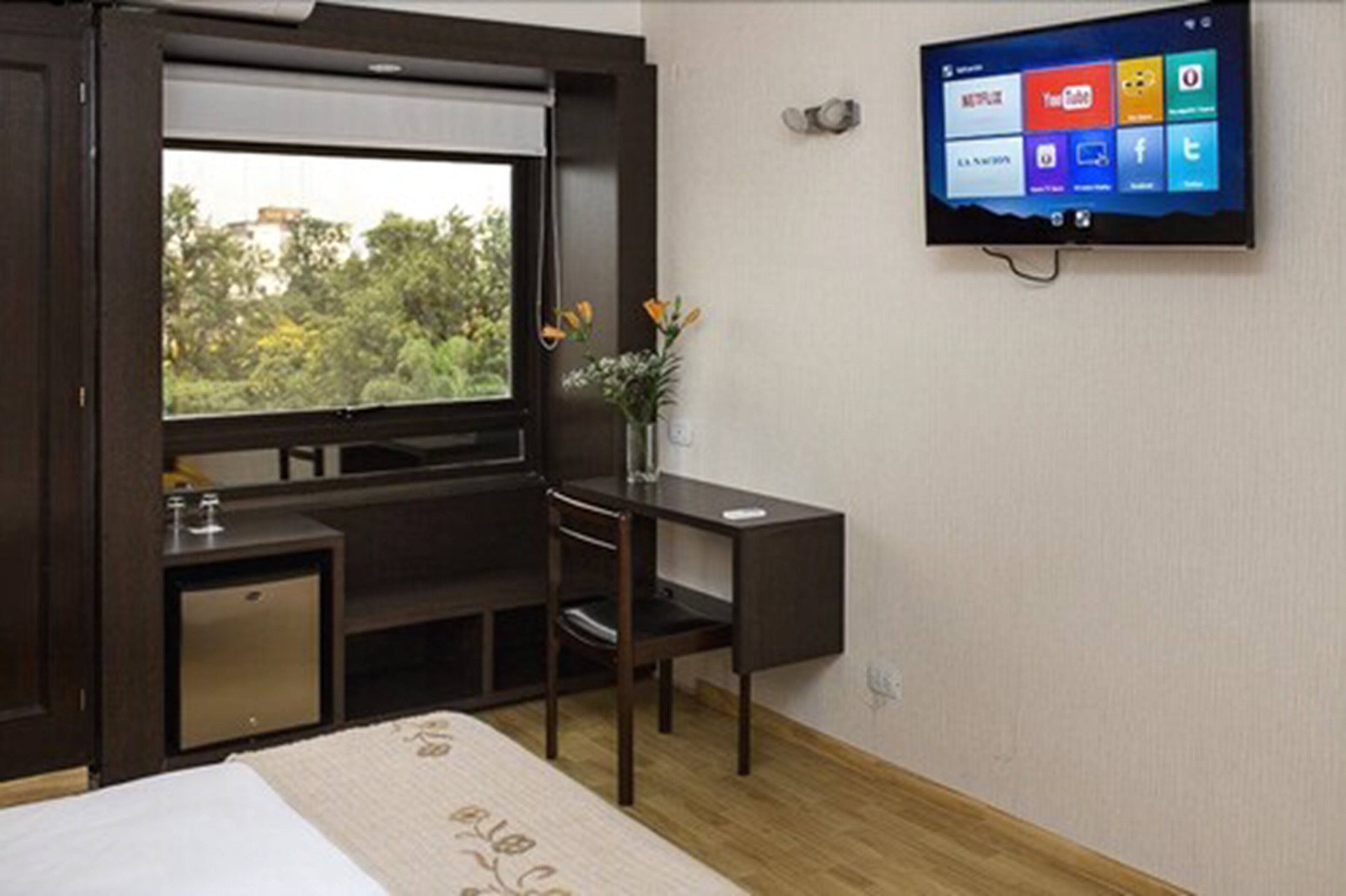 A hotel room with a bed, a desk, a chair, a small cabinet, a TV mounted on the wall, and a window with a view of greenery.