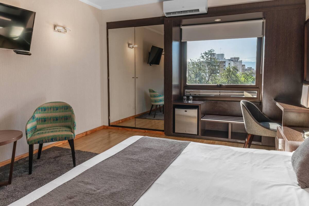 A hotel room with a bed, a desk, a chair, a TV, and a view of buildings through a window.