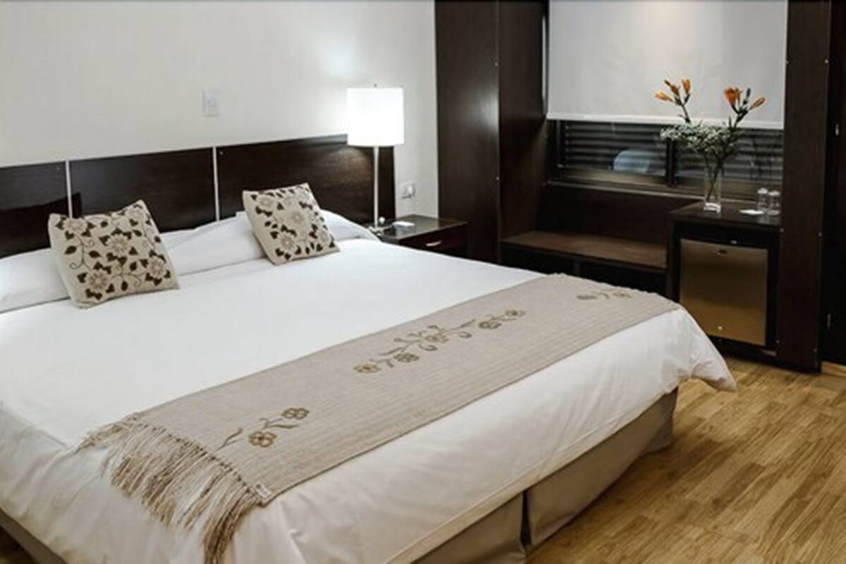 A hotel room with a large bed, a bedside table with a lamp, a small seating area, and a window with flowers.