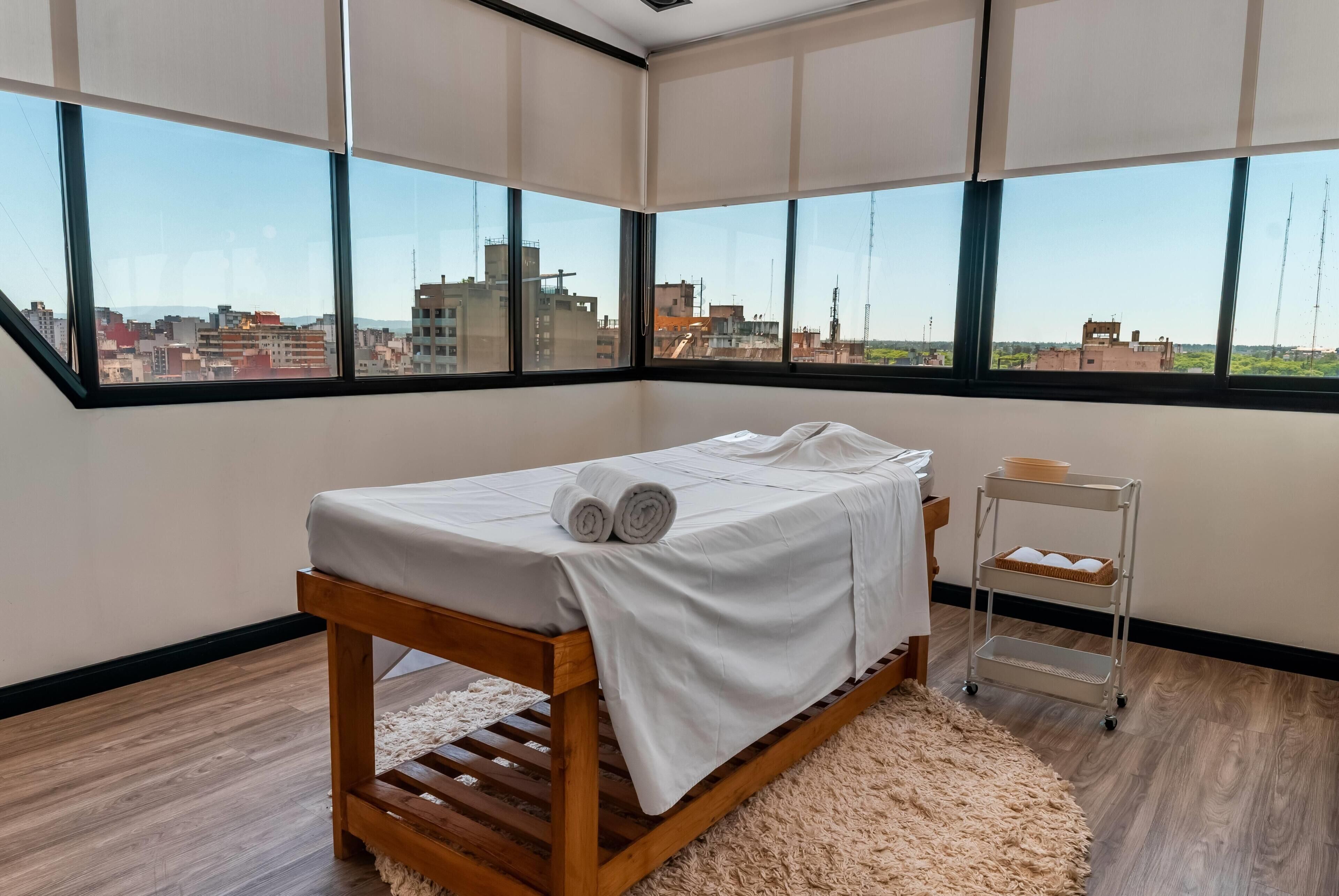 A room with a bed, a small table, a cart, and large windows with a city view.