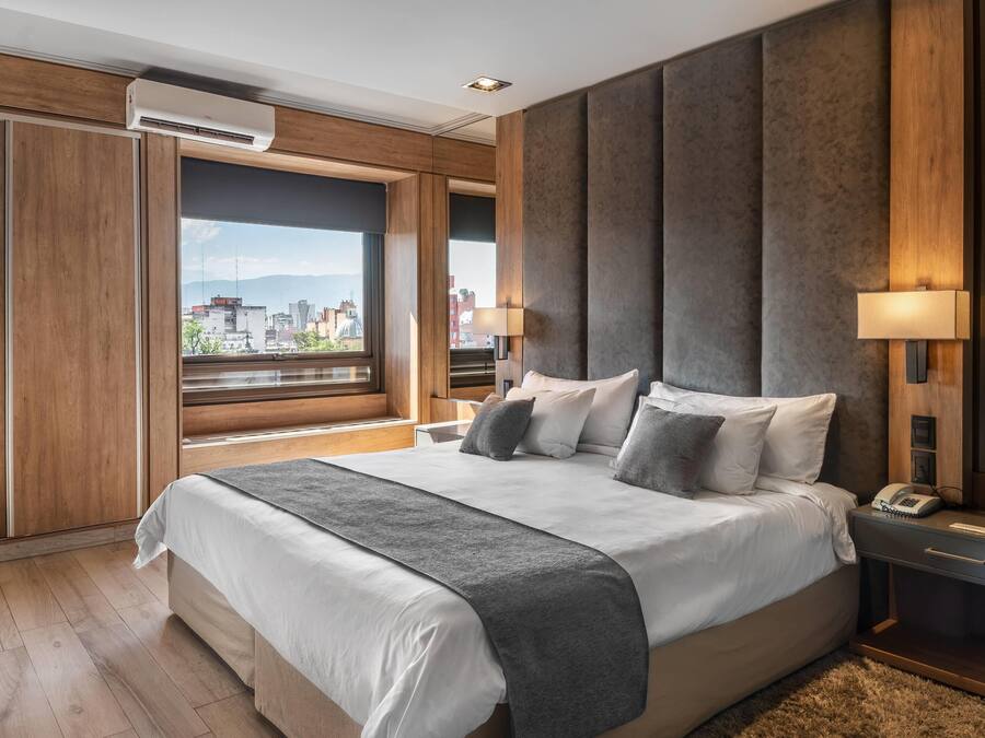 A modern hotel room with a large bed, a view of the city, a wooden wardrobe, and a desk with a lamp.