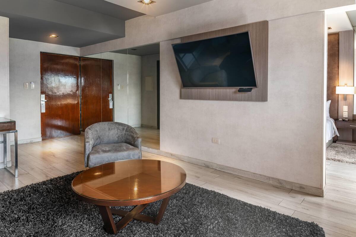 A modern living room with a wooden coffee table, a grey armchair, and a large flat-screen TV mounted on the wall.