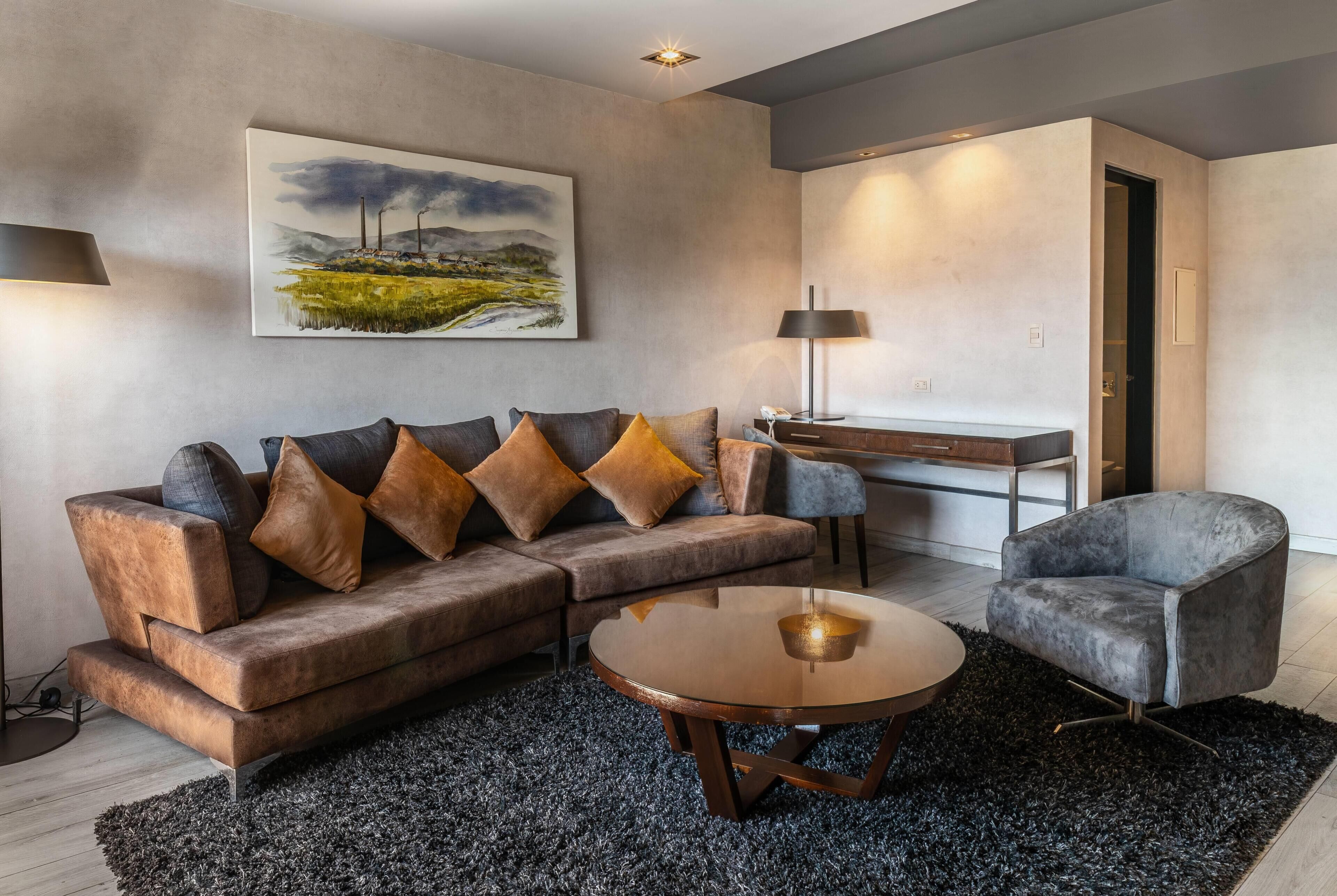 A modern living room with a sectional sofa, a round coffee table, a grey armchair, a painting on the wall, and a desk with a lamp.