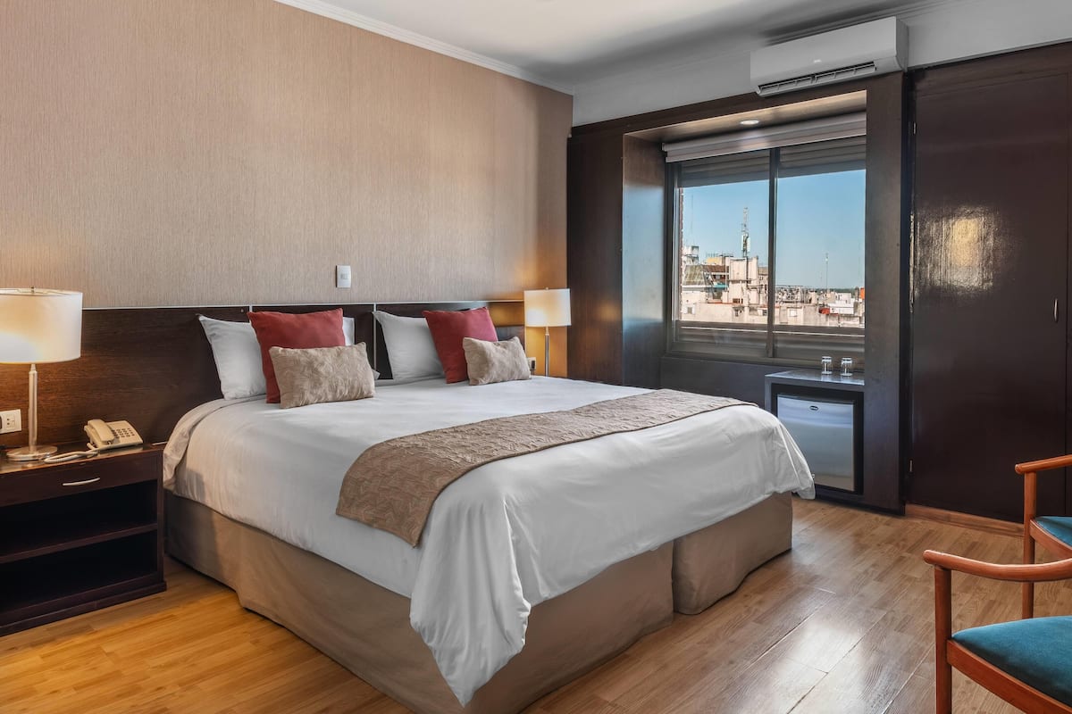 A hotel room with a large bed, bedside lamps, a nightstand with a phone, a small table with a lamp, a chair, and a view of the city through a large window.