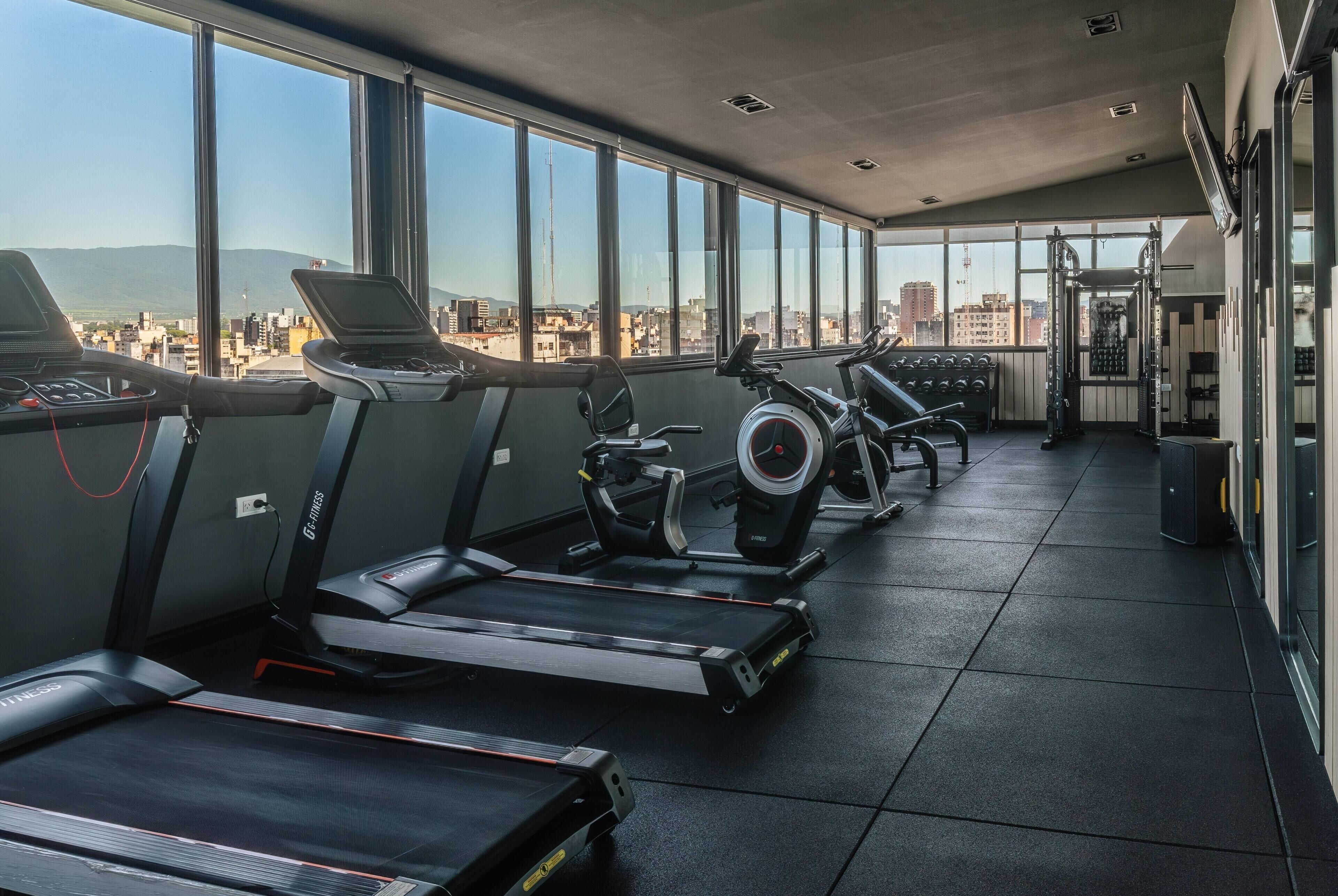 Un gimnasio con cintas de correr, una bicicleta estática y una máquina de pesas, con grandes ventanales que ofrecen vistas a la ciudad.