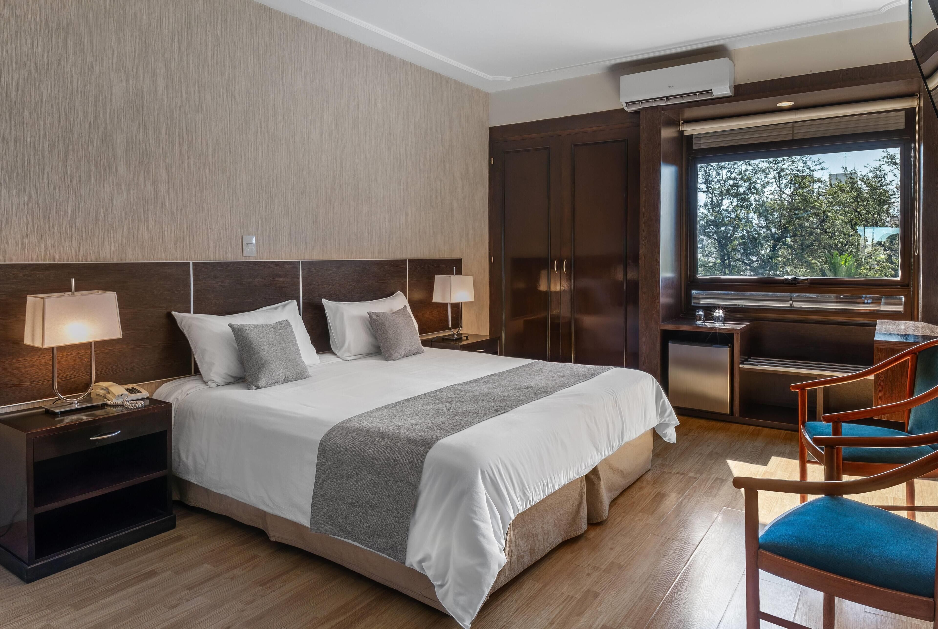 A hotel room with a large bed, bedside lamps, a desk, and a view of trees through the window.