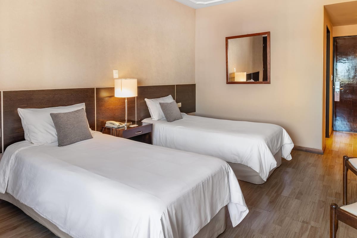 A hotel room with two beds, a bedside table with a lamp, a phone, and a mirror on the wall.