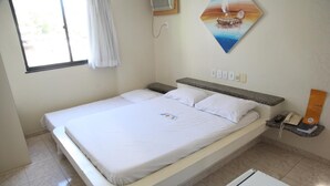 Ein Zimmer mit einem Einzelbett, einer Bettdecke in Weiß, einem Nachttisch mit Telefon und einem kleinen, orangefarbenen Gegenstand, einem Bild an der Wand und einem Fenster mit Vorhängen.