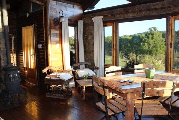 A rustic wooden interior with a large table, chairs, a fireplace, and a view of the outdoors.