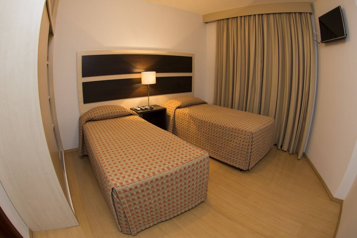 Habitación de hotel con dos camas individuales, una mesita de noche con lámpara, un televisor montado en la pared y una ventana con cortinas.