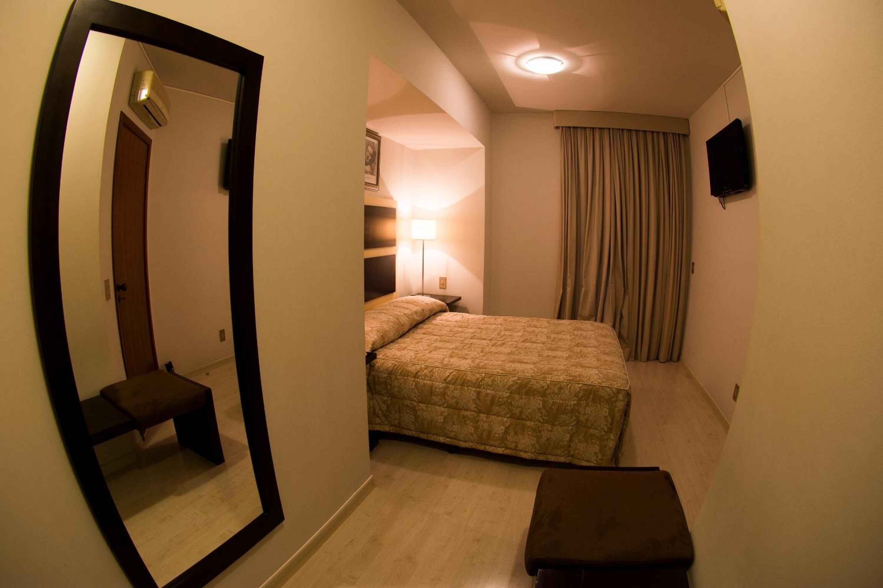 Quarto de hotel com cama, mesa de cabeceira, espelho, abajur e televisão.