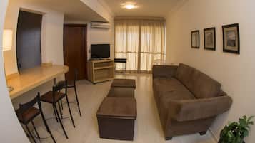 A living room with a brown sofa, a wooden coffee table, and a TV on a stand. There are two chairs and a small table.