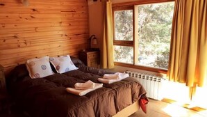 A wooden-paneled bedroom with a large window, a bed with brown bedding, and a nightstand with a lamp. - Apart Hotel Tierra Nativa (San Martín de los Andes)