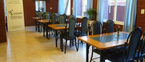 Un salón de conferencias con varias mesas y sillas, un cartel en la pared y ventanas con cortinas.
