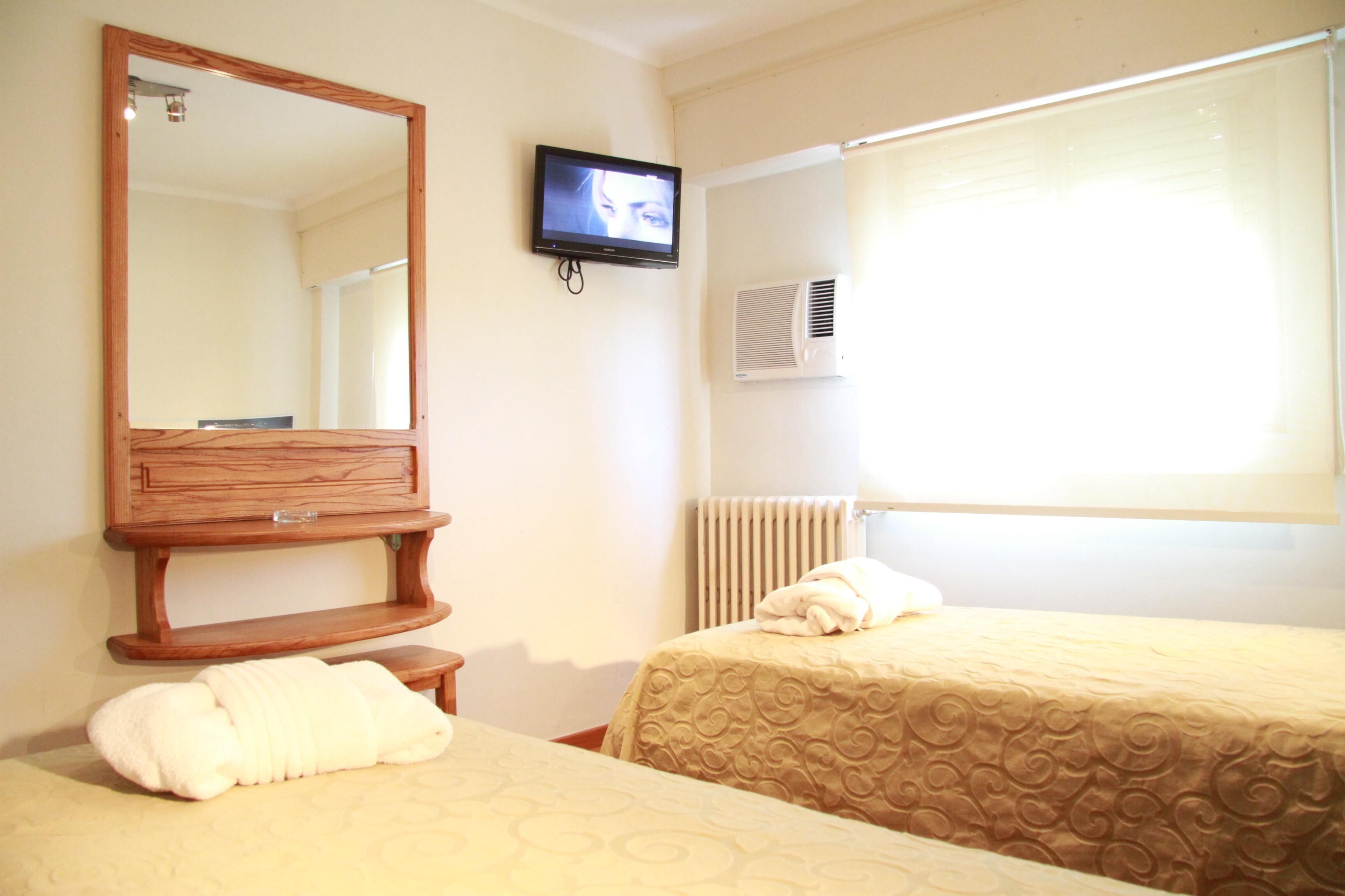 Habitación de hotel con dos camas, un televisor montado en la pared, un aparador de madera y una ventana con persianas.