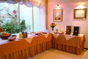 A breakfast buffet setup with a variety of food items and beverages, a vase with flowers, and framed pictures on the wall. - Kalton Hotel (San Rafael)