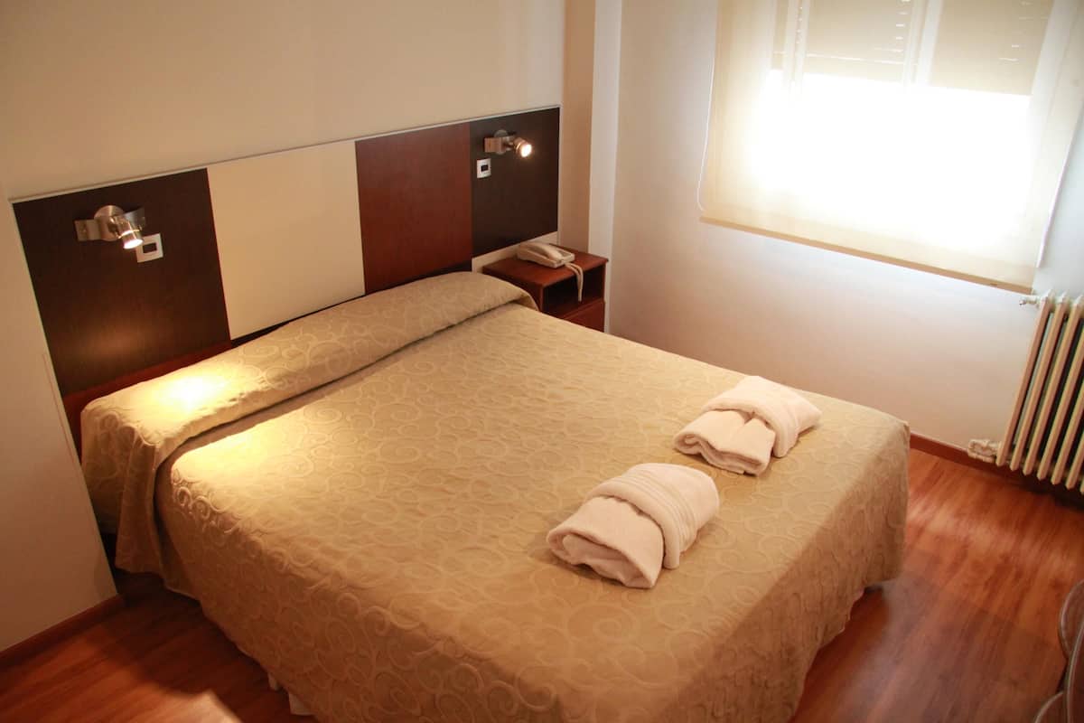 A double bed with a beige patterned bedspread and two folded towels, a bedside table with a phone, and a window with blinds.